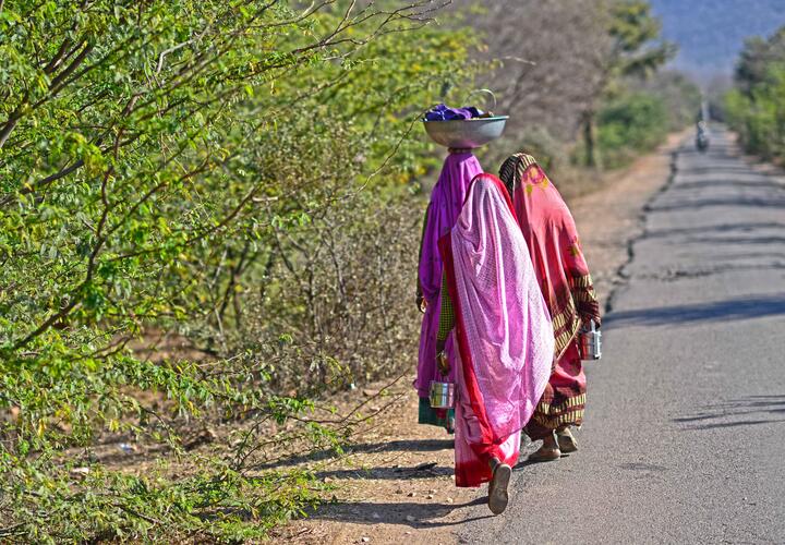 Photo of two women carrying baskets on their heads walking on the street. 